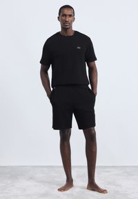 Homem está descalço em cima de um tapete, vestindo uma camisa de manga curta preta com logotipo e calções pretos, com as mãos nos bolsos, contra um fundo simples.