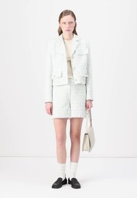Mėtų žalios spalvos tweed kostiumas, kuriame yra prigludusi striukė su kišenėmis ir mygtukais, derinama su atitinkamomis šortais ir loafer'iais, kartu su rankine.