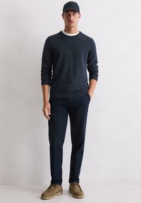 Granatowy sweter z prążkowanymi mankietami i dołem, biały okrągły dekolt, w zestawieniu z ciemnogranatowymi spodniami i jasnobrązowymi zamszowymi butami. Noszona czarna czapka.