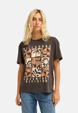 Blondine die een donker grafisch T-shirt met geometrische en natuurmotieven en een blauwe spijkerbroek draagt, staand tegen een effen achtergrond.