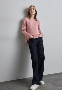 Rosa gerippter Pullover mit geometrischen Mustern, kombiniert mit dunkelblauen Schlaghosen. Das Model trägt weiße Sneakers und posiert vor einer grauen Wand.