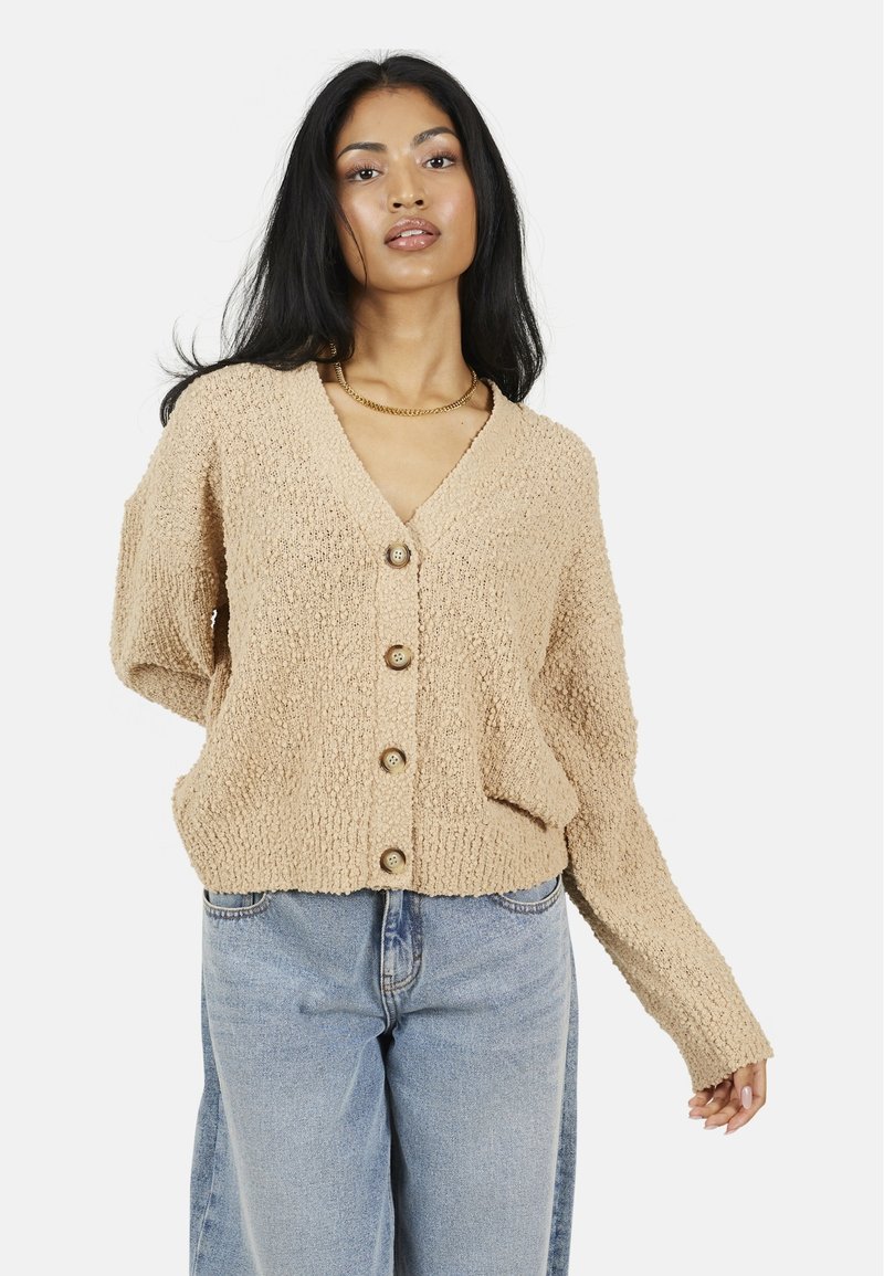 Cardigan beige texturé avec un col en V, cinq boutons en bois et des manches longues. Assorti à un pantalon large en jean bleu clair.