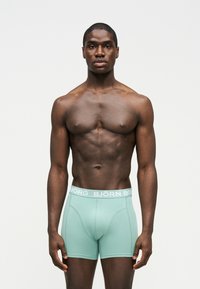 Άνδρας με γυμνό στήθος και μυώδη κορμό, φοράει ανοιχτό τιρκουάζ μπόξερ με λευκή μέση "BJÖRN BORG", στέκεται μπροστά σε απλό λευκό φόντο.