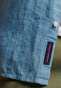 Camicia azzurra a maniche corte con bottoni, realizzata in tessuto testurizzato, con un piccolo etichetta nera con testo rosa sull'orlo.
