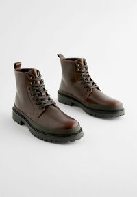 Botas de cuero marrón hasta el tobillo con suelas de goma negras, punteras redondas y cordones grises. Con detalles de costura y tiradores en la parte trasera.