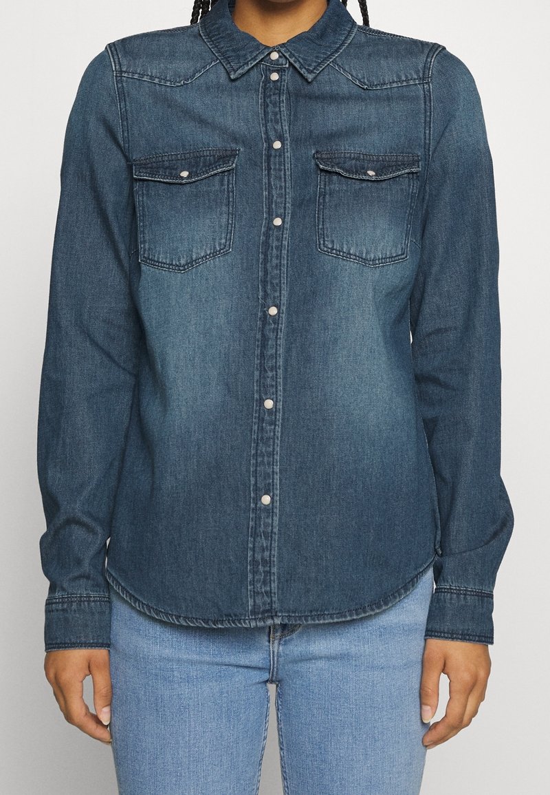 Personne portant une chemise en denim bleu foncé à manches longues avec des boutons-pression et deux poches sur la poitrine, associée à un jean bleu clair.