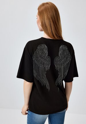 Femeie cu păr roșu purtând un tricou negru oversized cu un model cu aripi de înger albe punctate pe spate și blugi albaștri.
