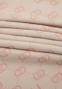 Tessuto beige con una consistenza morbida, caratterizzato da una leggera lucentezza. Decorato con un motivo a logo arancione chiaro ripetuto e dettagli di cucitura.