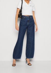 Blå vida jeans i denim, med framfickor, bältesloopar och hög midja. Kombineras med en kortärmad vit polo-shirt och svarta klackar.