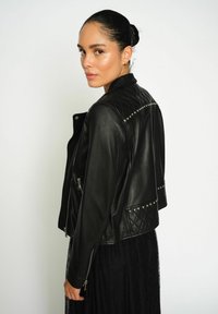 Moteris su tamsiais plaukais surišta į kuodą, vilkinti juodą odinį švarką su gumbuotais ir siuvinėtais akcentais virš juodos nėrinių sijono, žiūrinti per petį.