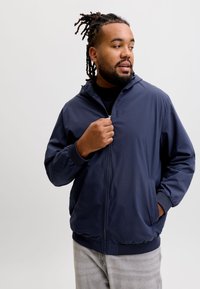 Homme avec des dreadlocks portant une veste zippée bleu marine et un pantalon gris clair, regardant sur le côté avec une main touchant la fermeture éclair.