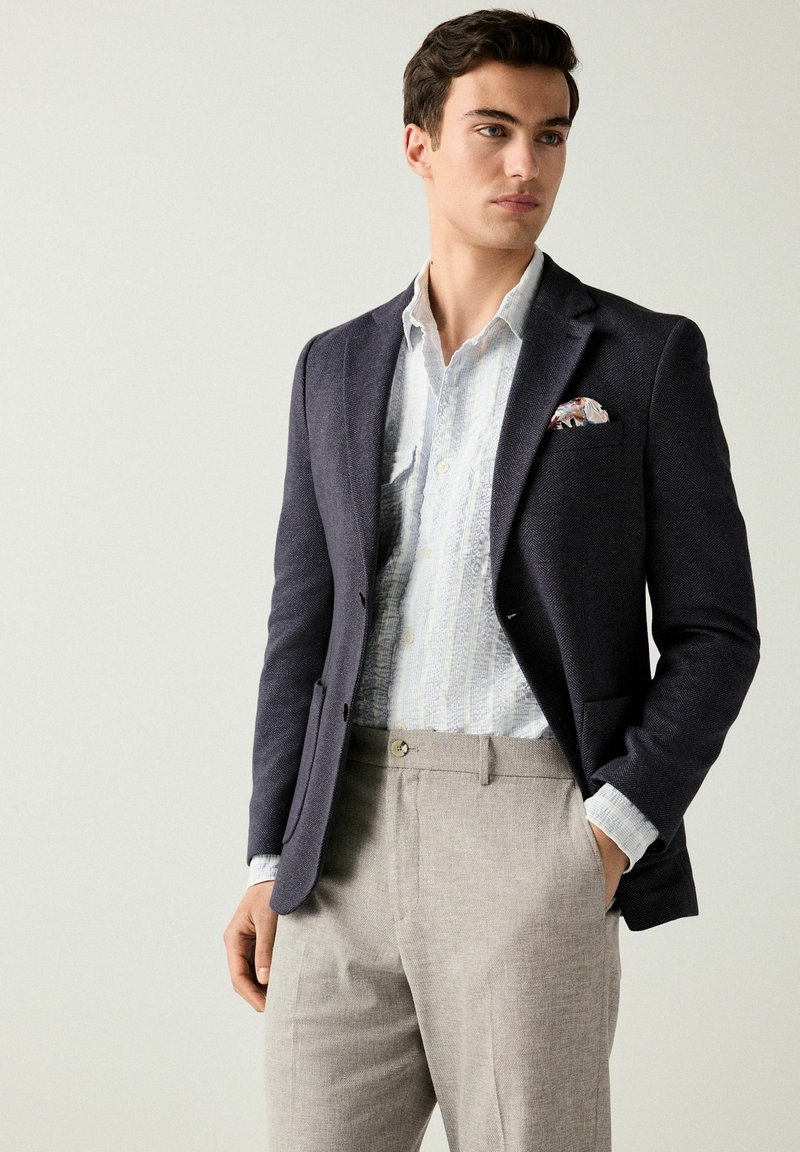 Donker marineblauw colbert over een witte gestructureerde shirt, lichtgrijze broek, met een geprint pochet in de borstzak van het colbert.