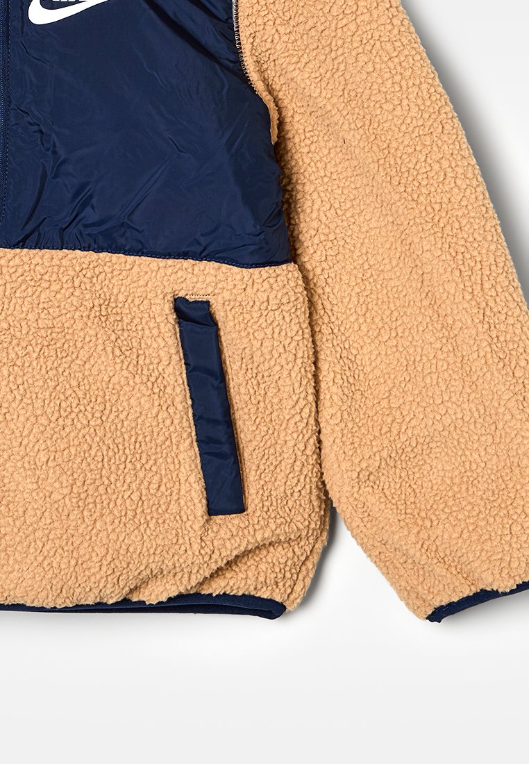 Veste en polaire avec un corps texturé beige et un haut en nylon bleu marine, poche zippée et logo de la marque sur la poitrine pour un détail supplémentaire.
