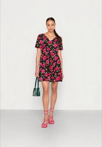 Vestido floral com fundo preto, mangas curtas e decote em V. Padrões de flores rosa e verde. A modelo segura uma bolsa verde e usa sandálias rosas.