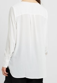 Blusa bianca a maniche lunghe in un tessuto leggero e trasparente. Presenta una piega sulla schiena e polsini con bottoni, con un orlo arrotondato e una trama morbida.