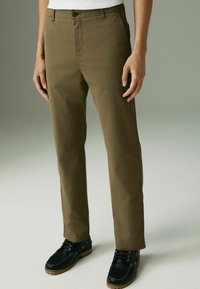 Pantalons en coton kaki avec une coupe droit, fermeture à bouton et poches latérales. Associés à des chaussures noires avec une semelle texturée.