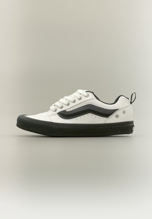 Biały sneaker z czarnymi akcentami, z gumową podeszwą, sznurowaniem oraz teksturowaną cholewką z zamszu. Minimalistyczny design z detalem logo.