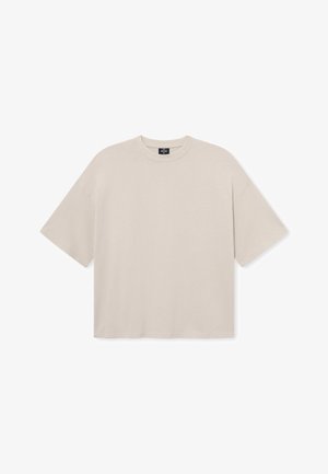 Beżowy, oversize t-shirt z miękkiego materiału, z okrągłym dekoltem i krótkimi rękawami, na modelce z kręconymi włosami.