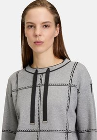 Šedá mikina s kapucí se vzorem mřížky, černými šňůrkami a kontrastním šitím. Má kulatý výstřih a strukturovanou tkaninu.