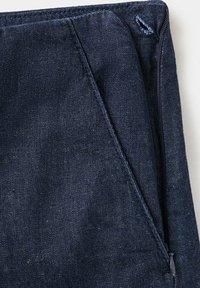 Gros plan d'un tissu en denim bleu foncé avec une fermeture éclair latérale partiellement dézippée et une boutonnière cousue sur la ceinture.