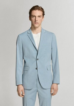 Giovane uomo con indosso un abito azzurro chiaro e una camicia bianca, in piedi con un'espressione neutra contro uno sfondo chiaro e uniforme.