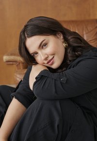 Jeune femme aux cheveux foncés et boucles d'oreilles en or assise, serrant ses genoux, souriant doucement, portant une tenue noire avec un fauteuil en cuir marron en arrière-plan.