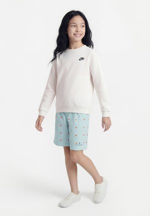 Fille aux longs cheveux foncés portant un sweat-shirt blanc Nike, un short bleu clair à pois rouges et des baskets blanches, debout et souriante.
