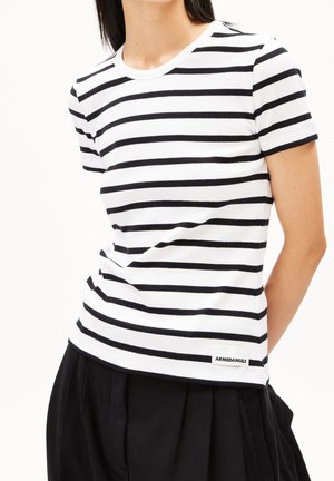 Kobieta ubrana w białą koszulkę z krótkim rękawem z czarnymi poziomymi paskami oraz czarne spodnie, z metką "ARMEDANGELS" blisko krawędzi.