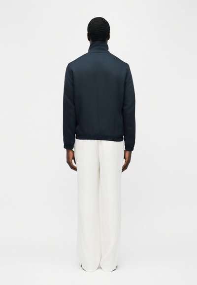 Homme debout, dos tourné, portant une veste bleu marine foncé à col haut et un pantalon blanc ample, sur un fond clair uni.