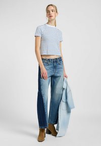 Randig vit och blå kort t-shirt i kombination med vida tvåtoners denimjeans. Modellen håller en ljusblå skjorta. Bruna klackstövlar med bred klack.