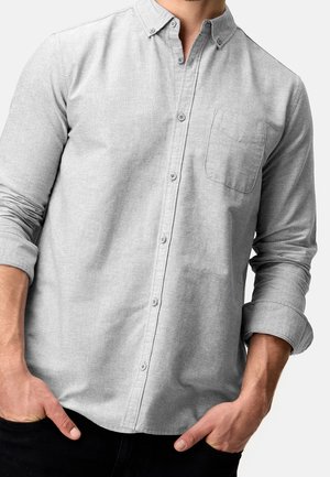 Homme portant une chemise gris clair boutonnée, manches retroussées, les mains dans les poches d’un pantalon noir, sur un fond uni.
