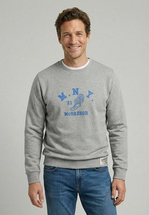 Lachende man met kort bruin haar, die een lichtgrijze trui draagt met een blauwe opdruk "M.N.Y. '21 McGREGOR" en een afbeelding van een gevleugelde hardloopschoen, en een blauwe spijkerbroek.