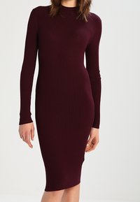 Robe en tricot côtelé bordeaux, manches longues, silhouette ajustée, longueur au genou, texture lisse, encolure ronde sans embellissements visibles.