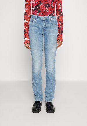 Λεπτά γαλάζια τζιν παντελόνια φορεμένα με μαύρες loafers και ένα μακρυμάνικο κόκκινο μπλουζάκι με λουλούδια, στανταρισμένα σε μια απλή ανοιχτόχρωμη φόντο.