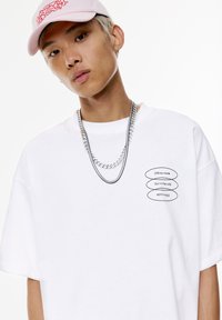 T-shirt bianco oversize con etichette nere con la scritta "Modalità offline", "In mezzo alla natura" e collane a catena argentata sovrapposte. Cappellino rosa chiaro.
