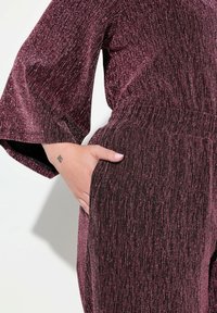 Burgundy jumpsuit med glitrende tekstur, brede ærmer og elastisk talje. Har en sidelomme på hoften.