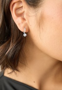 Pendiente de aro de plata con una piedra preciosa redonda y brillante. Presenta una superficie texturizada y un cierre seguro. Lleva puesto una modelo en la oreja.