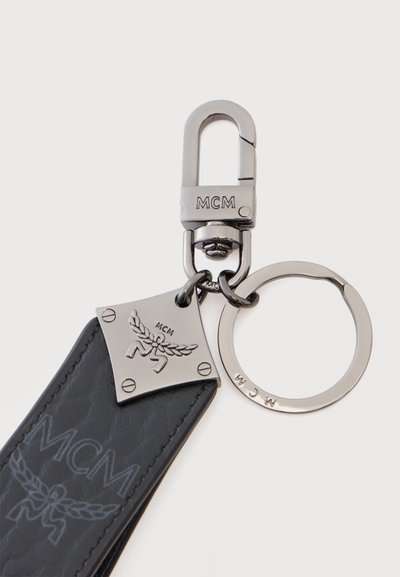 Porte-clés métallique avec logo MCM, sangle en cuir noir et fermoir sur fond clair.