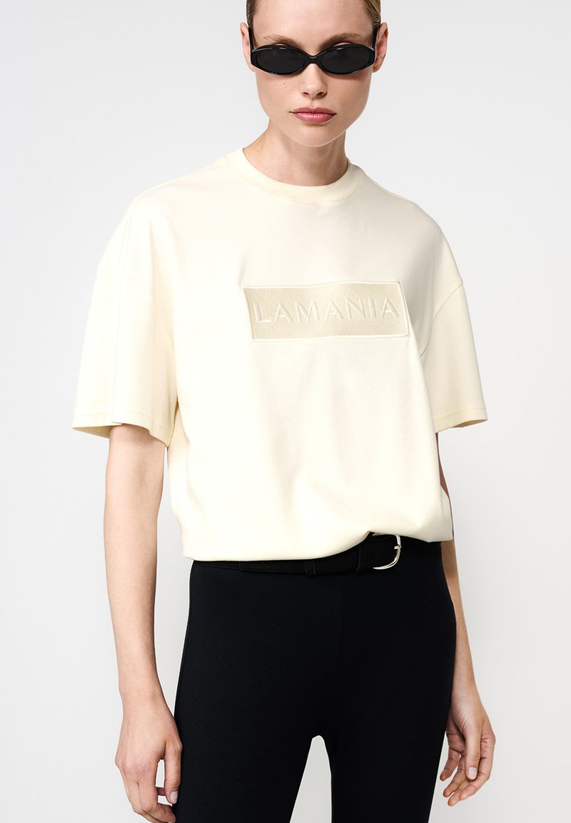 Moteris, vilkinti kreminę per didelę T-shirt su "LAMANIA" aplikacija, juodais priglundančiais džinsais, juodu diržu ir plonais juodais akinių nuo saulės rėmeliais, prieš neutralų foną.