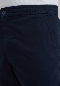 Pantalon bleu marine avec une texture lisse, taille élastique et fermeture à un bouton. Comprend des poches latérales et un détail de couture subtil.