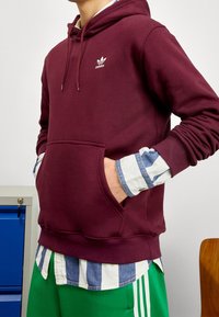 Maroon hoodie i bomullsmaterial med en framficka och vitt Adidas-logotyp. Under den syns en blå och vit randig skjorta.