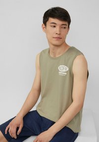 Olivgrünes ärmelloses T-Shirt mit weißem grafischem Text, kombiniert mit dunkelblauen Shorts. Baumwollmischung, lockere Passform, schlichtes Design.
