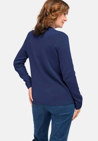 Vrouw met schouderlang golvend haar, gekleed in een marineblauwe trui en blauwe jeans, staande met haar rug naar de camera, terwijl ze opzij kijkt.