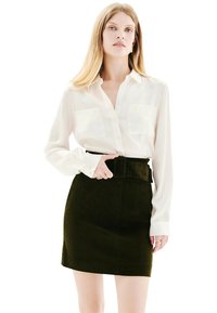 Jeune femme portant une chemise blanche à boutons rentrée dans une jupe noire taille haute avec une large ceinture, se tenant avec une main sur la ceinture.