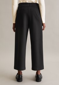 Pantalons larges noirs avec une finition lisse, présentant une taille haute, une fermeture éclair et des poches latérales. Portés avec des chaussures noires.