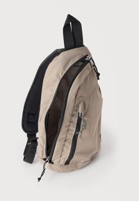 Kompakter beiger Sling-Rucksack mit schwarzem, gepolstertem Schultergurt, zwei Reißverschlussfächern und schwarzem-gelbem Kordelzug.