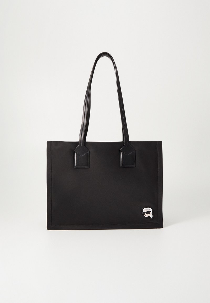 Sac cabas rectangulaire noir avec de longues anses en cuir et un petit patch cartoon représentant une personne avec des lunettes de soleil dans le coin inférieur droit.