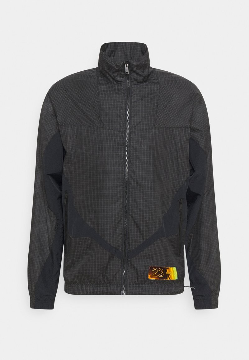Black jacket with a grid pattern, a high collar, zip closure, and contrasting fabric panels. Features a colored logo patch on the hem.