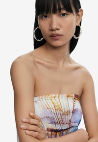 Õlavarreta kleit valgel taustal, millel on abstraktsed oranžid ja sinised mustrid. Mudel kannab suuri hõbedasi rõngas kõrvarõngaid ja hõbesõrmust.