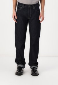 Jeans de mezclilla azul oscuro con corte recto, que presentan costuras en contraste, diseño de cinco bolsillos y dobladillos remangados. Combinados con zapatos negros.